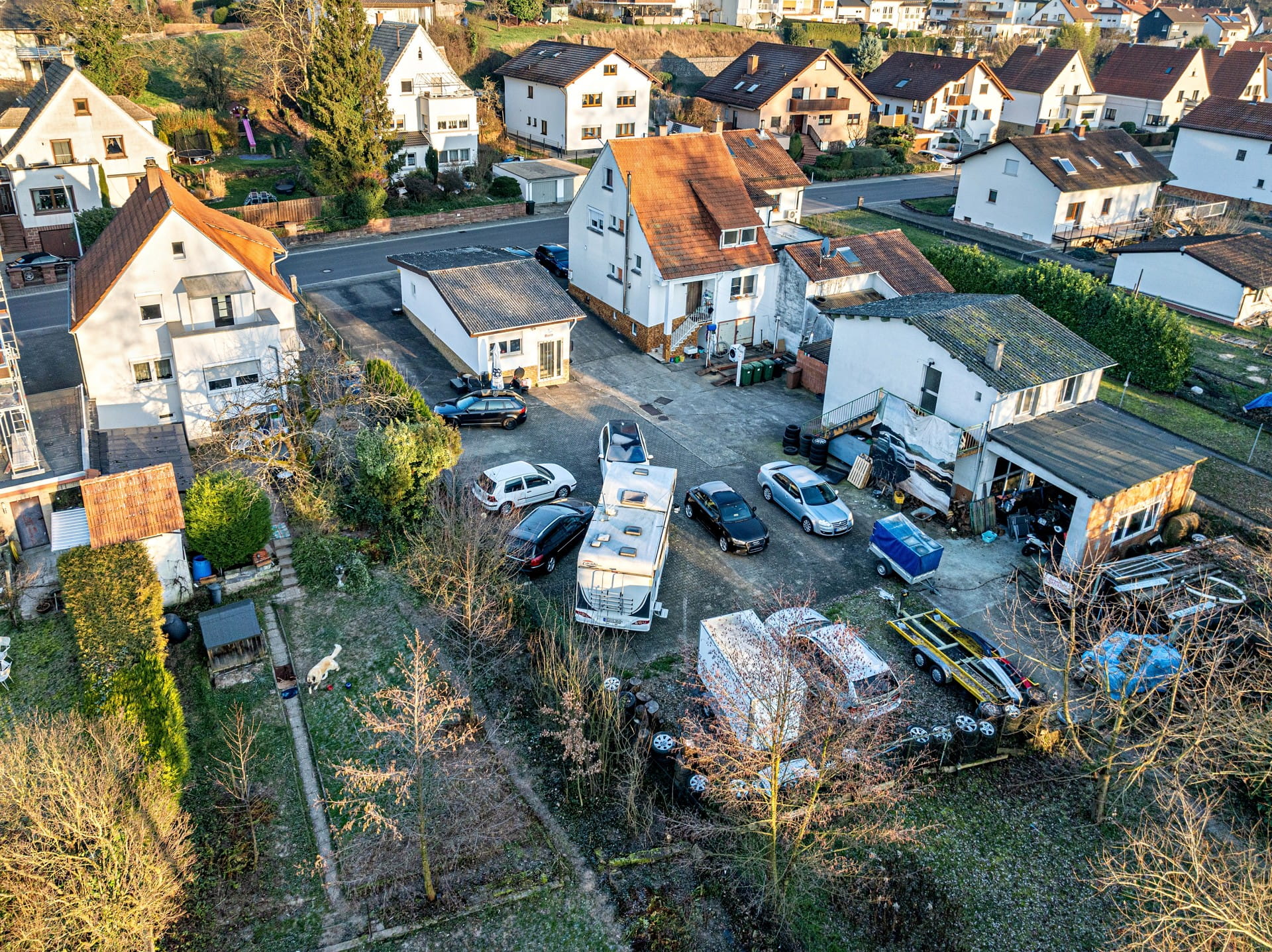 Eine Drohnenaufnahme zeigt ein weitläufiges, aber stark zugestelltes Grundstück in einem Wohngebiet. Der große befestigte Innenhof zwischen den Bestandsgebäuden wird als Abstellfläche für zahlreiche Autos, ein Wohnmobil, Anhänger, Reifen und diversen Hausrat genutzt. Auch der angrenzende Gartenbereich im Vordergrund mit kahlen Bäumen wirkt unstrukturiert.